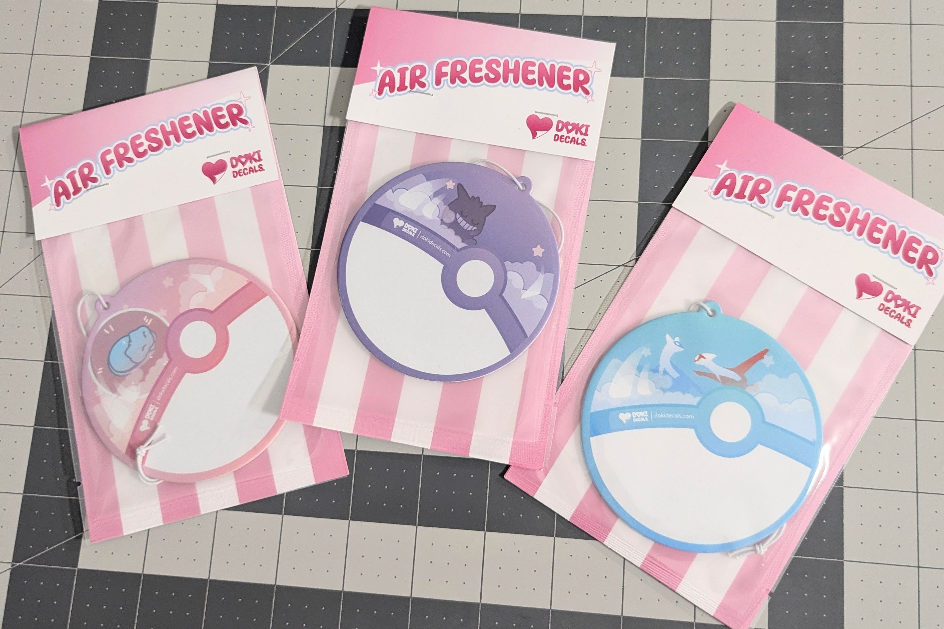 Three air freshener packages with pink and white striped design on a grid background