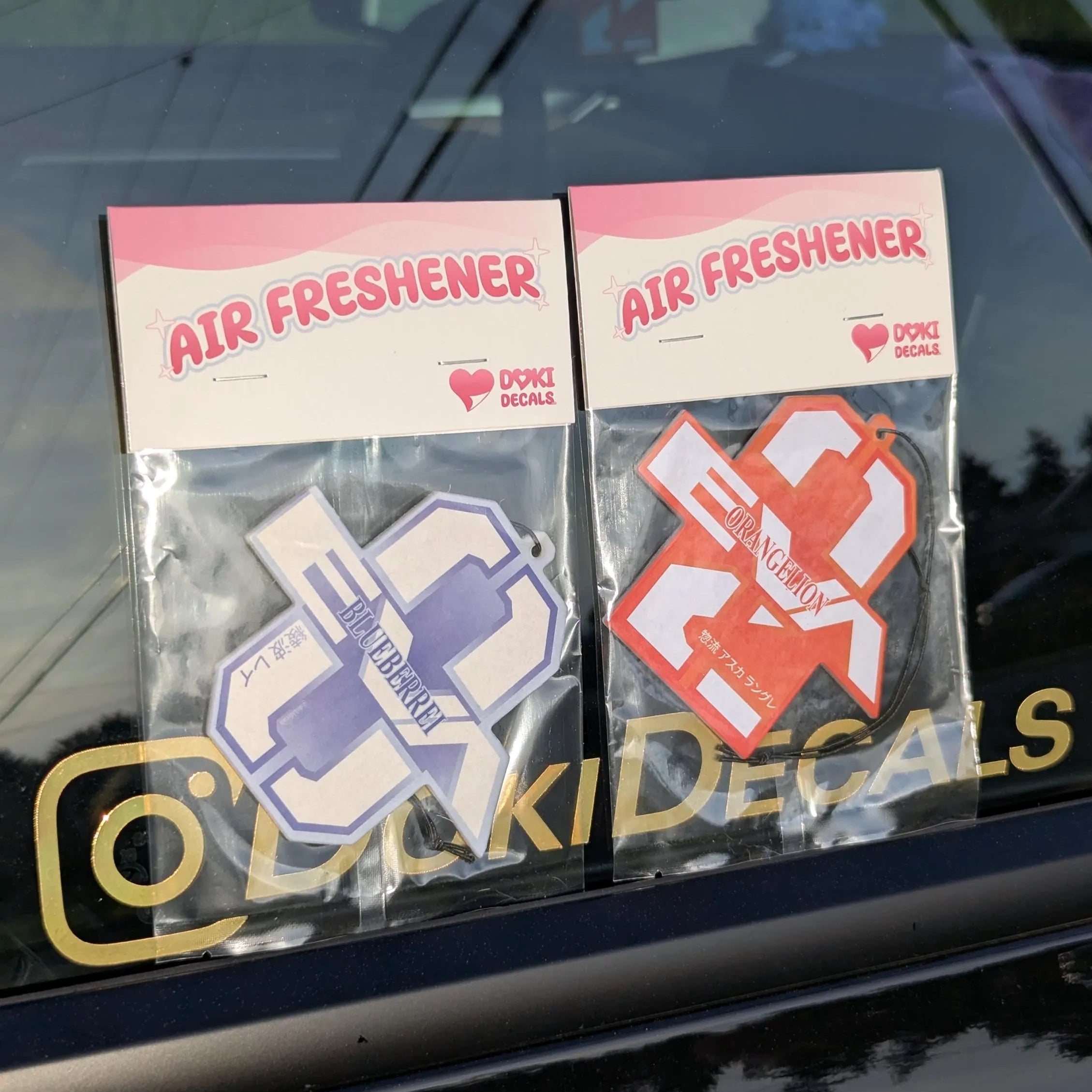 Two air freshener packages with decal designs on a car window, featuring the brand 'Doki Decals'.